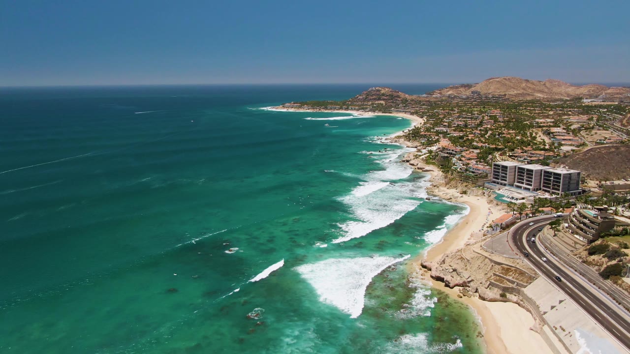 Costa Azul Beach aerial frame with a large white wave fan in the lower center, clear green-blue water offshore, coastal buildings to the right, and no visible people.