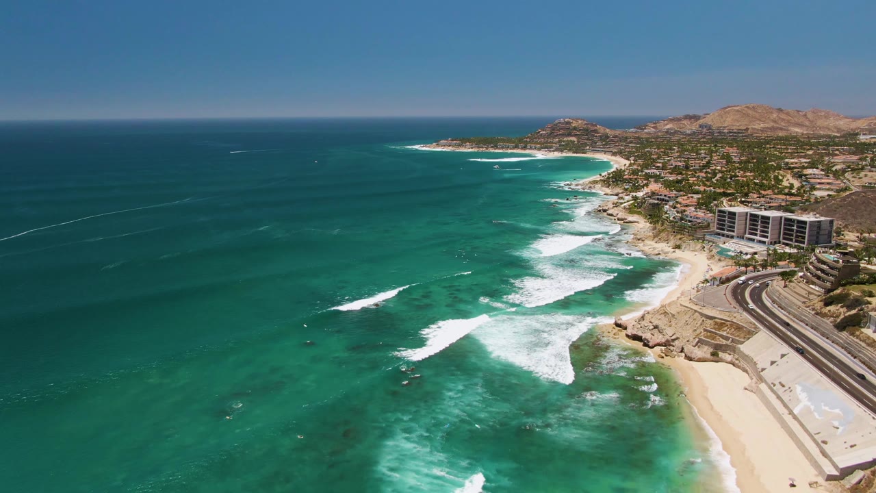 Aerial frame at Costa Azul Beach in Los Cabos showing turquoise water, white surf, a sandy shore, a coastal road, shoreline buildings, tan hills, and no visible people.