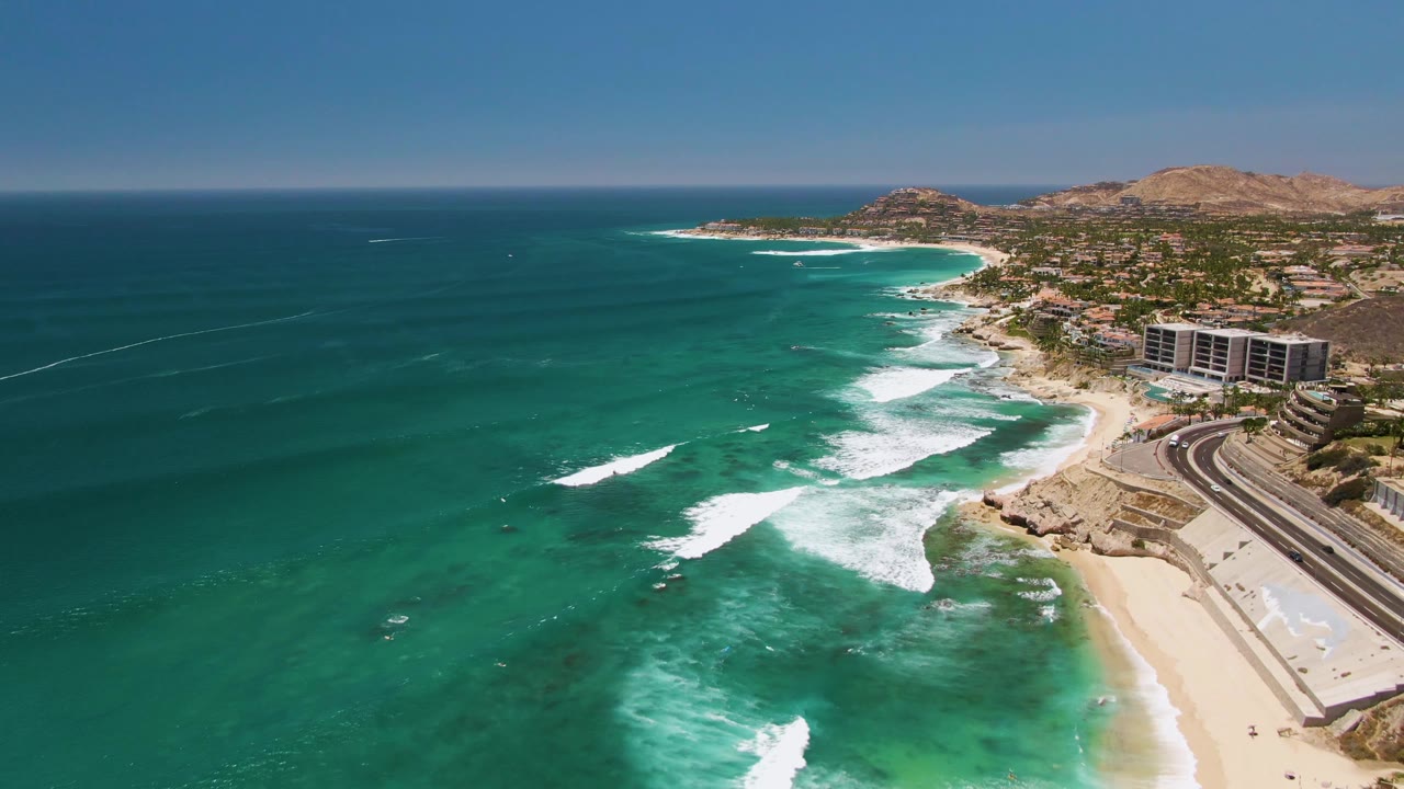 Aerial frame at Costa Azul Beach in Los Cabos with a larger white wave band crossing the nearshore water, beach and road at right, coastal buildings inland, and no visible people.