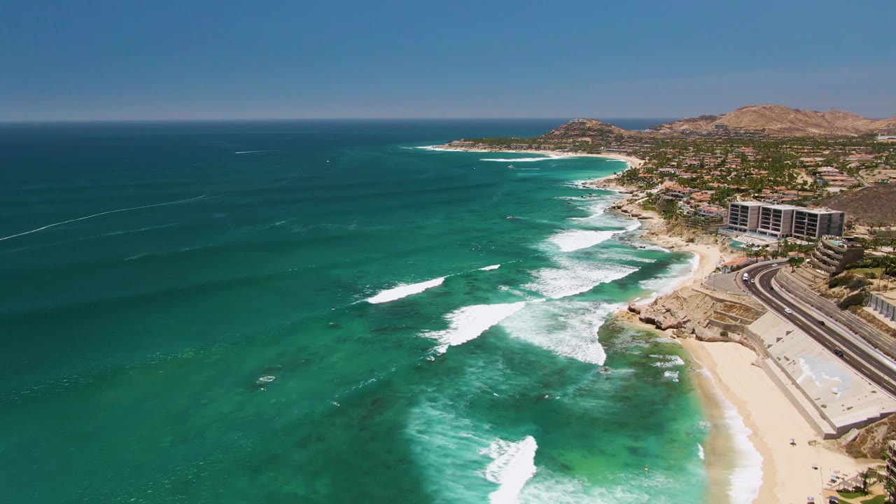 Aerial frame at Costa Azul Beach in Los Cabos where the coastline sits farther right, the open ocean fills more of the frame, white wave foam crosses the middle, and no visible people appear.