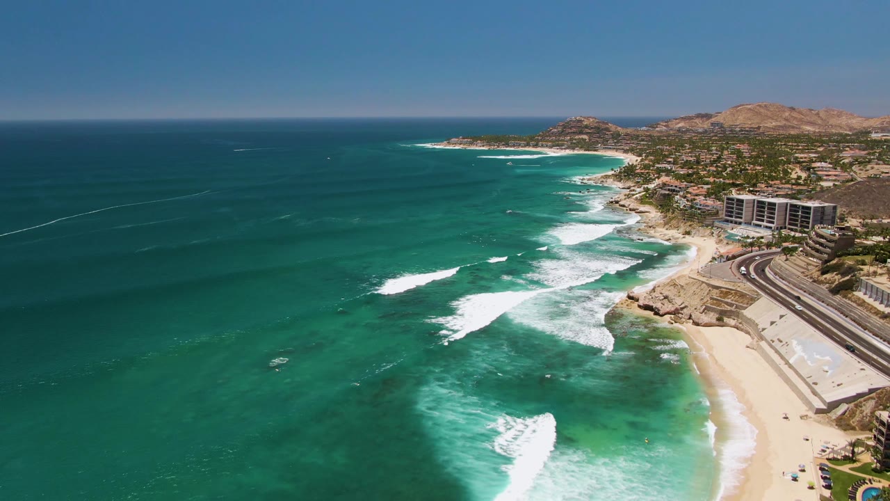 Drone view looking along a beach where white waves break beside coastal buildings.