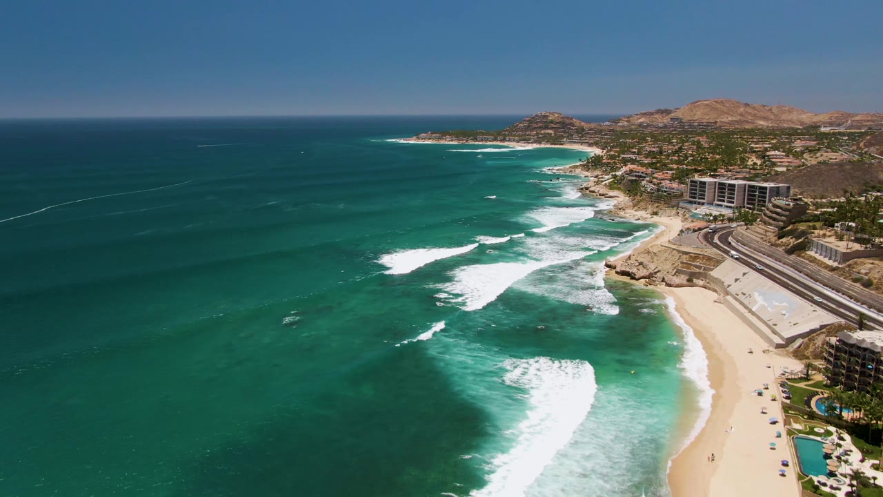 Opening aerial frame over Costa Azul Beach in Los Cabos, with turquoise water on the left, white surf bands reaching the sand, resort buildings on the right, and tiny beachgoers visible.