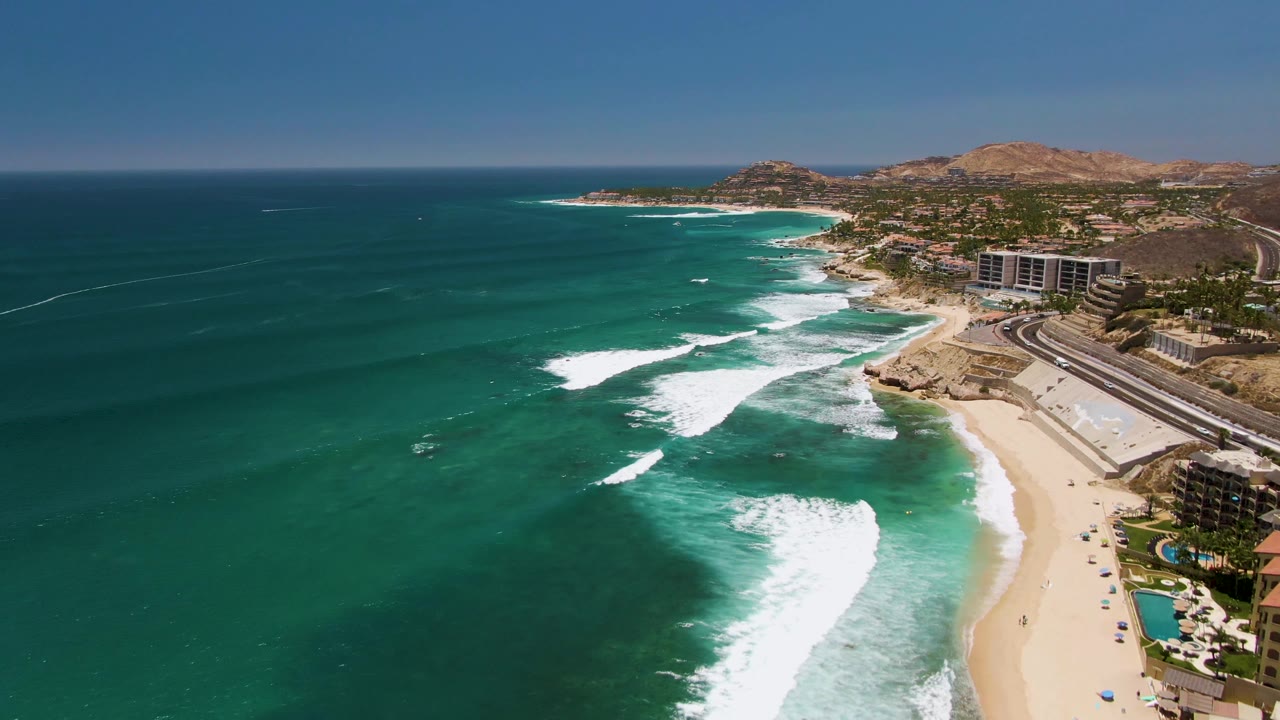 Wide drone view of white surf bands moving toward a sandy beach with buildings and hills beyond.