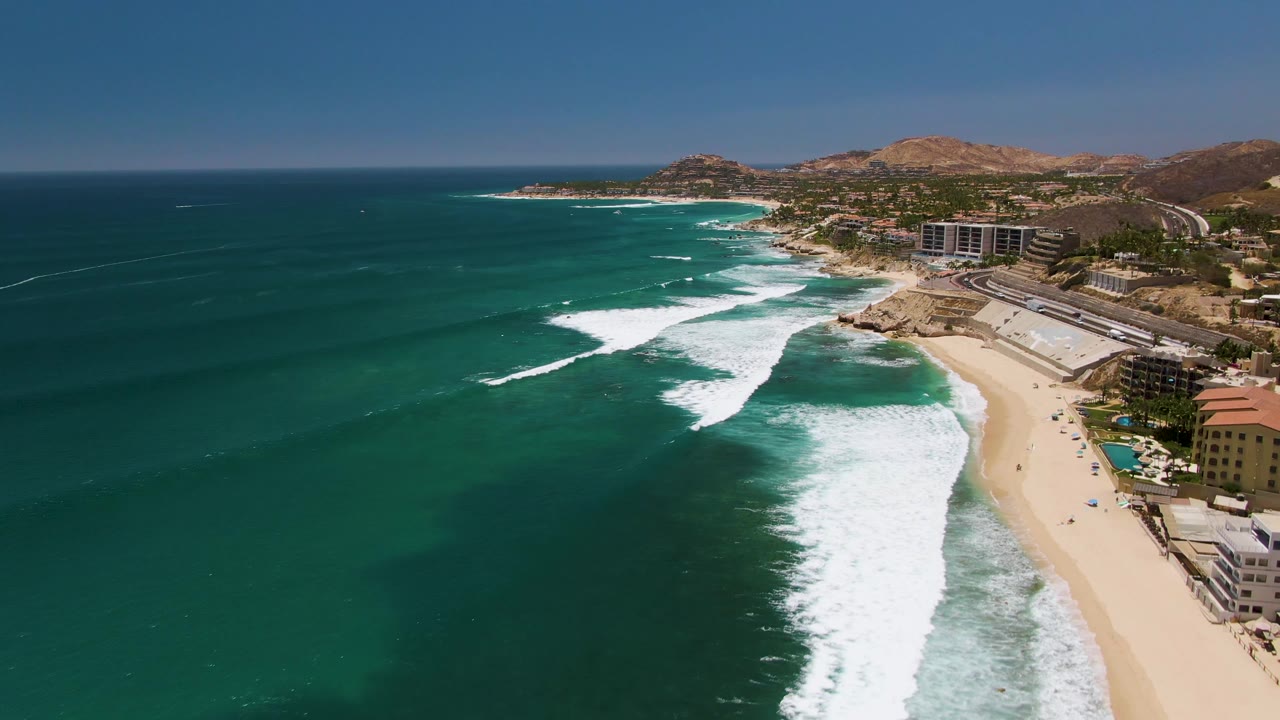 At 0:00, an aerial view over Costa Azul Beach in Los Cabos shows turquoise water, white surf bands, a sandy shoreline, coastal buildings, and dry hills beyond the beach.