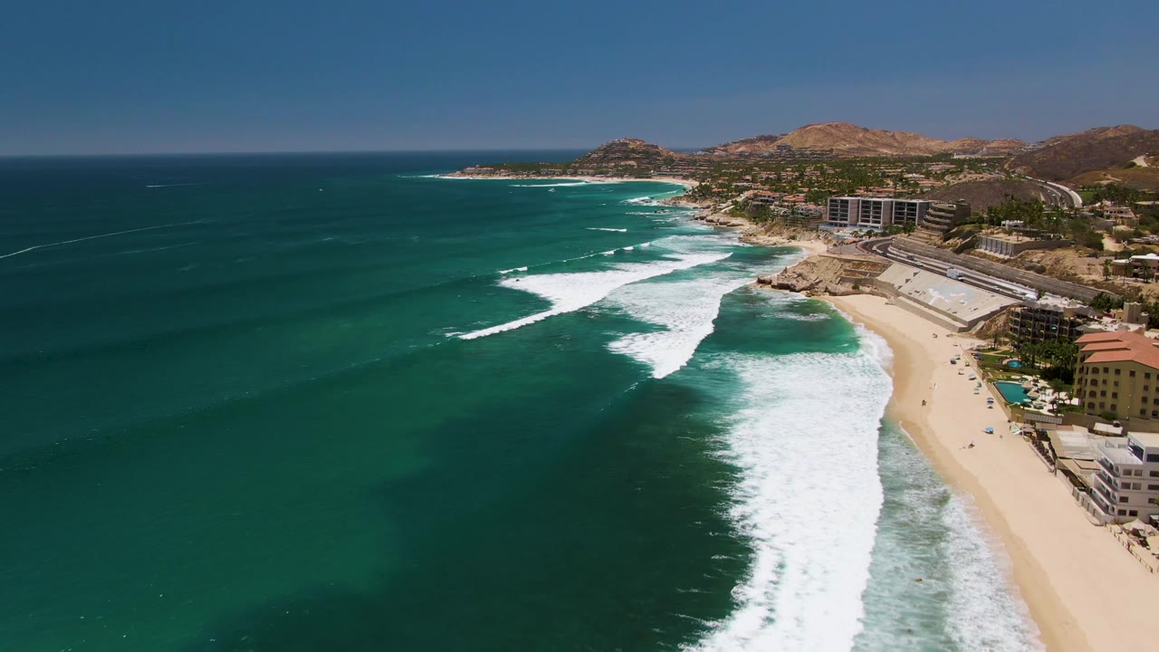 White surf lines break across turquoise water beside a sandy beach and coastal buildings.