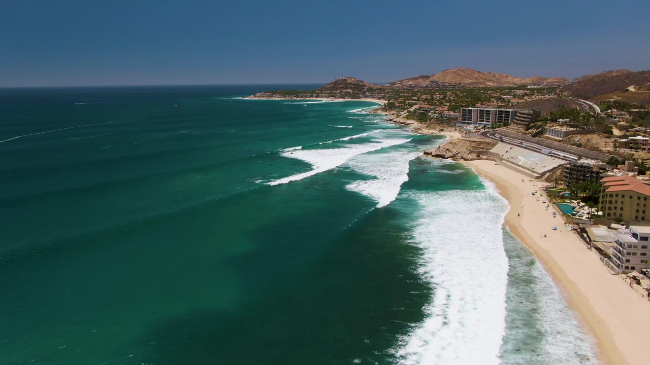 At 0:02, the aerial frame shows a broad sweep of Costa Azul Beach with open ocean on the left, a bright foam path near shore, buildings at right, and hills behind them.