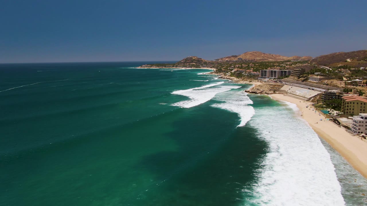 Aerial frame over Costa Azul Beach in Los Cabos showing turquoise water, white surf curling onto sand, coastal buildings at right, distant dry hills, and tiny visible people near the shore.