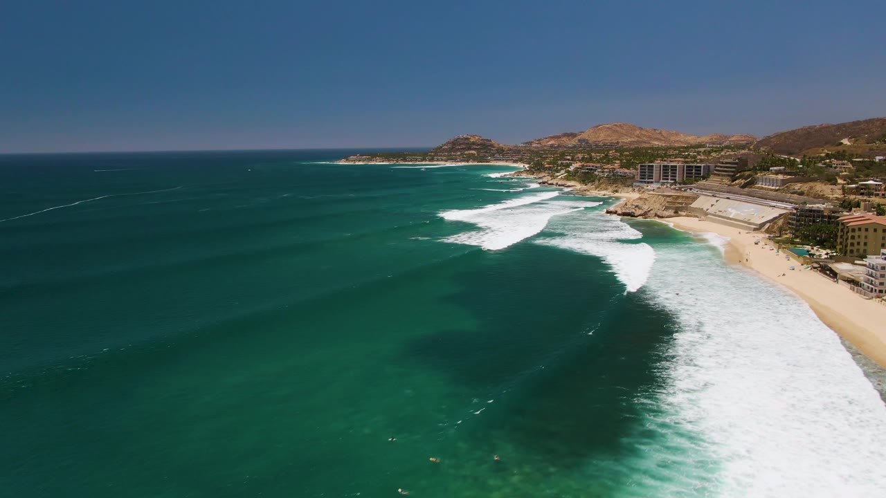 Aerial view over Costa Azul Beach with open turquoise water across the foreground, broken white wave lines near the sand, coastal buildings at right, arid hills behind, and small people visible as dots in the water.