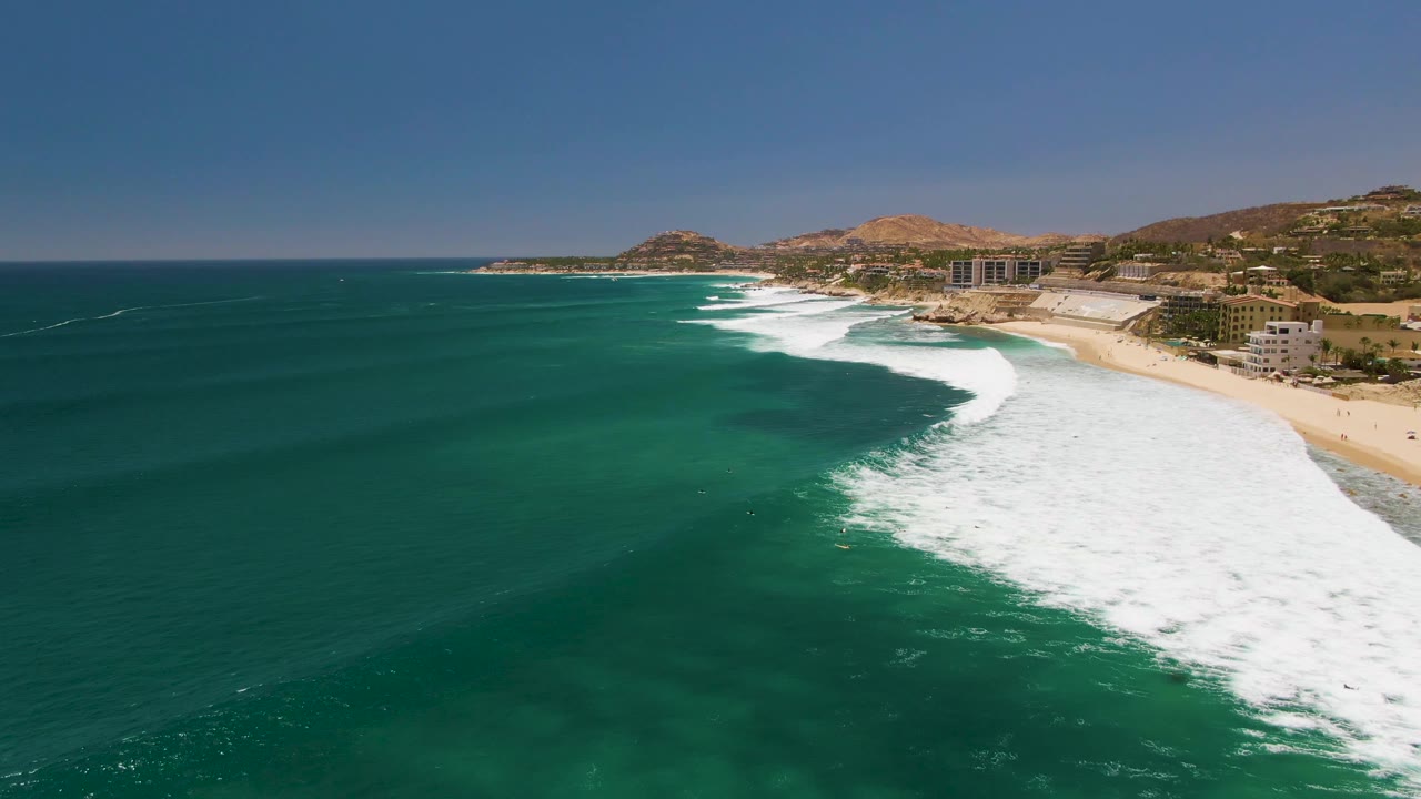 At Costa Azul Beach in Los Cabos, turquoise water fills the lower frame as a broad band of white surf breaks toward sand, with hillside buildings in the distance and no visible people.