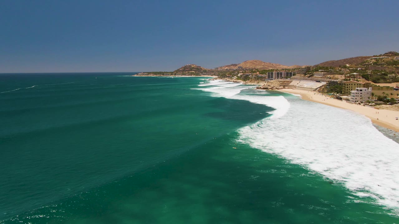 At Costa Azul Beach in Los Cabos, the surf band has advanced across more of the right side of the frame, with clear turquoise water in front, sand and buildings behind, and no visible people.
