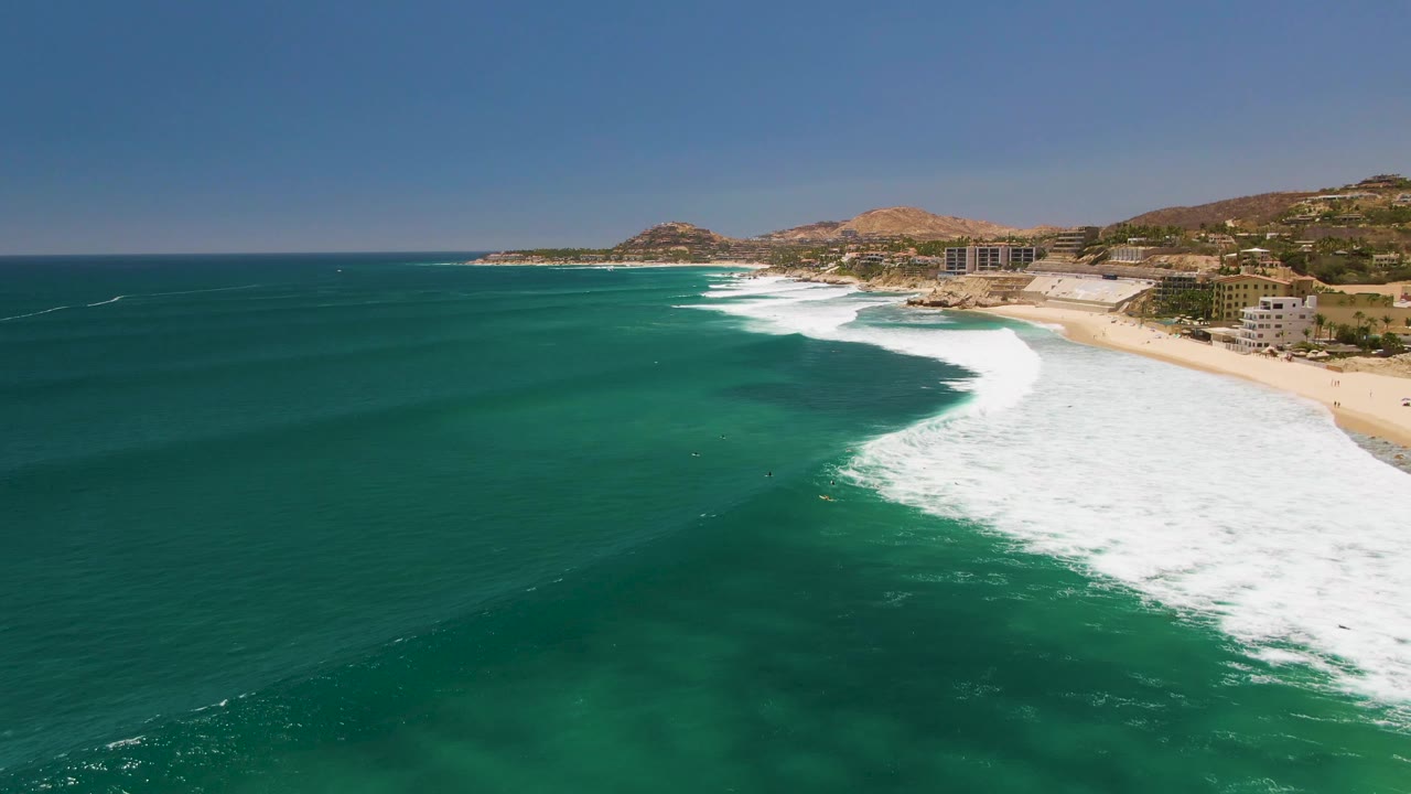 At Costa Azul Beach in Los Cabos, green-blue ocean occupies most of the frame while a bright white wave breaks close to the sandy shore, backed by low buildings and hills with no visible people.