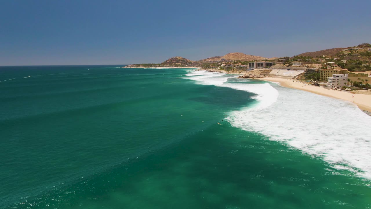 Aerial coastline view with dark turquoise water, foamy surf, sandy beach, buildings, and hills.