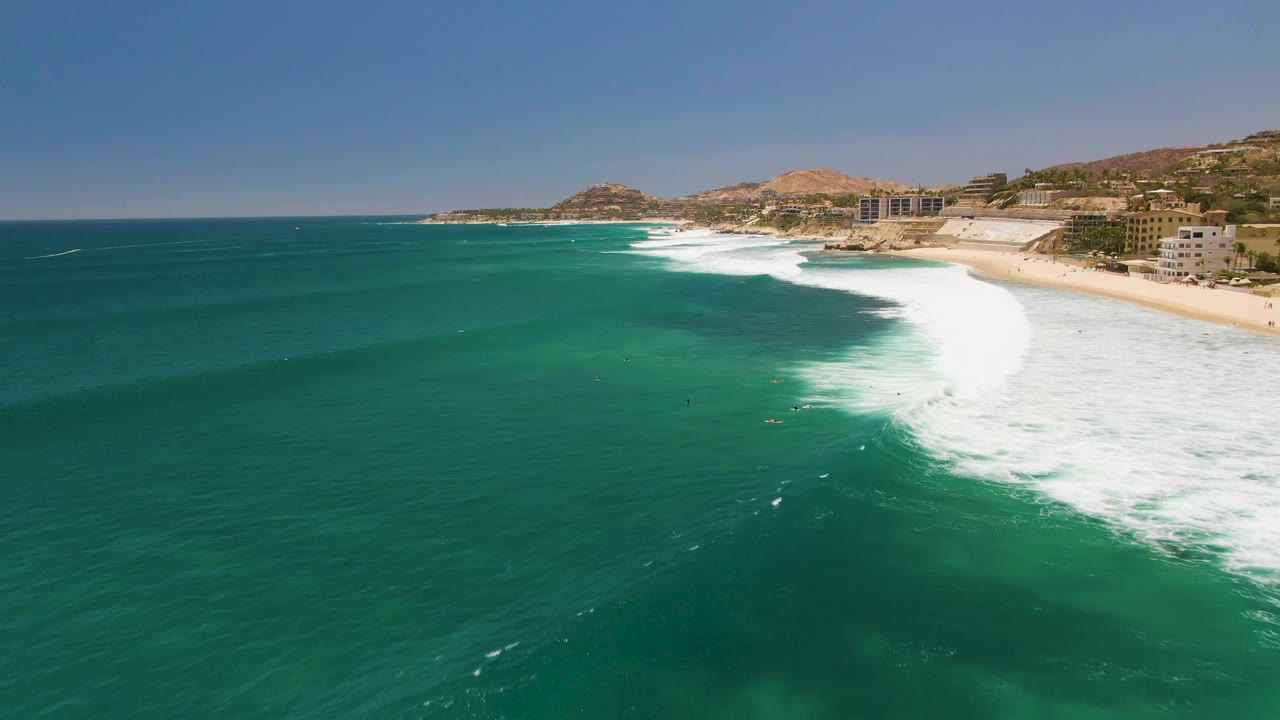 Opening aerial frame over Costa Azul Beach in Los Cabos, Mexico, with green-blue ocean, a white breaking wave near the sandy shore, small surfers in the water, coastal buildings, hills, and blue sky.