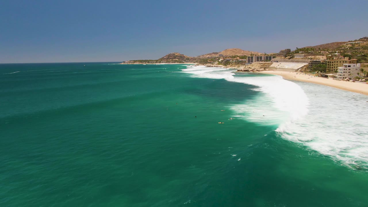 Turquoise ocean and white surf breaking toward a sandy shoreline with small surfers visible.