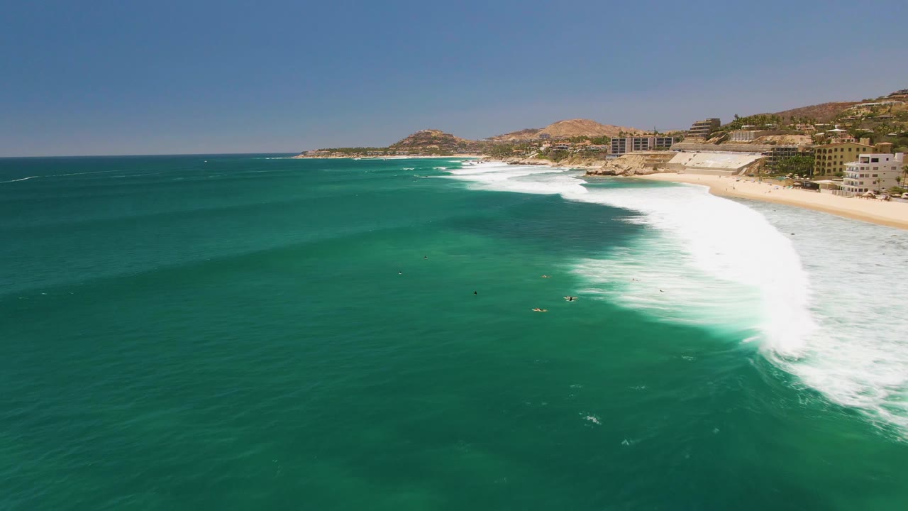 Mid-clip aerial frame at Costa Azul Beach with a broad white foam section on the right, darker green ocean across the foreground, small surfers near the break, beach buildings, brown hills, and blue sky.