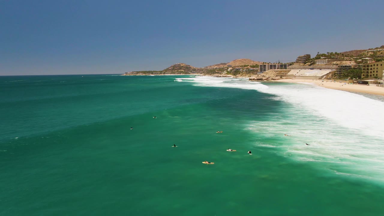 Wide aerial view of surfers and swimmers in turquoise water beside the sandy shore at Costa Azul Beach in Los Cabos, with white surf and coastal buildings in the distance.