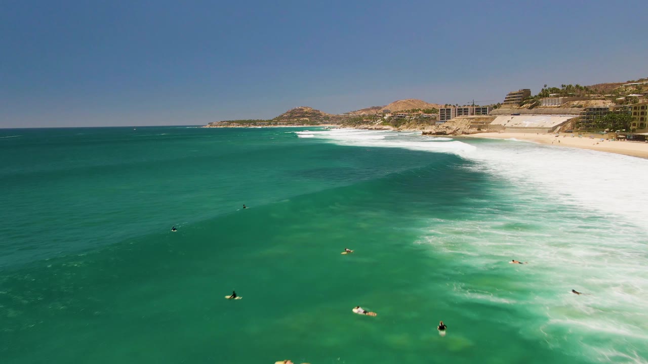 Costa Azul Beach in Los Cabos appears from above with scattered water users, a bright white surf band, sandy beach, coastal buildings, and dry hills beyond the shoreline.