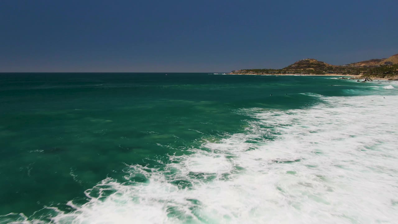Aerial view of turquoise ocean at Costa Azul Beach Los Cabos with white foam spreading across the right foreground, distant coastline and hills on the horizon, and no visible people.
