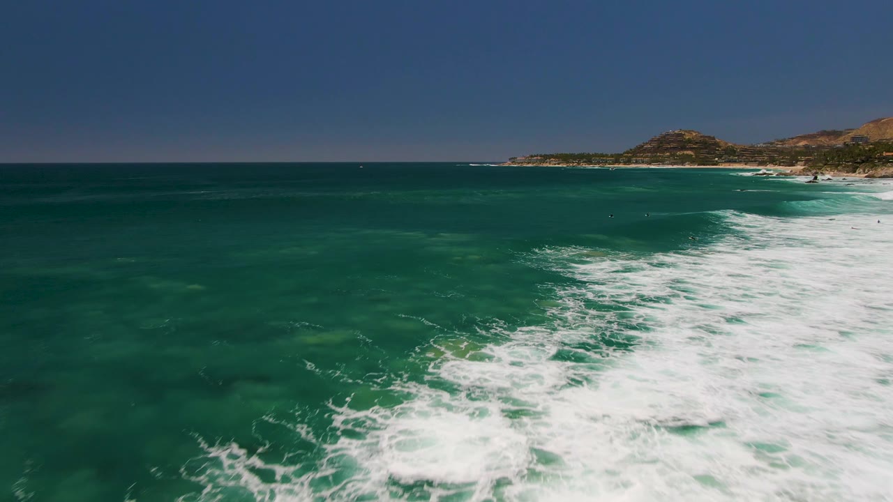 Aerial frame over green-blue water at Costa Azul Beach Los Cabos, with a broad white foam patch across the lower center-right and low hills beyond the coastline, with no visible people.