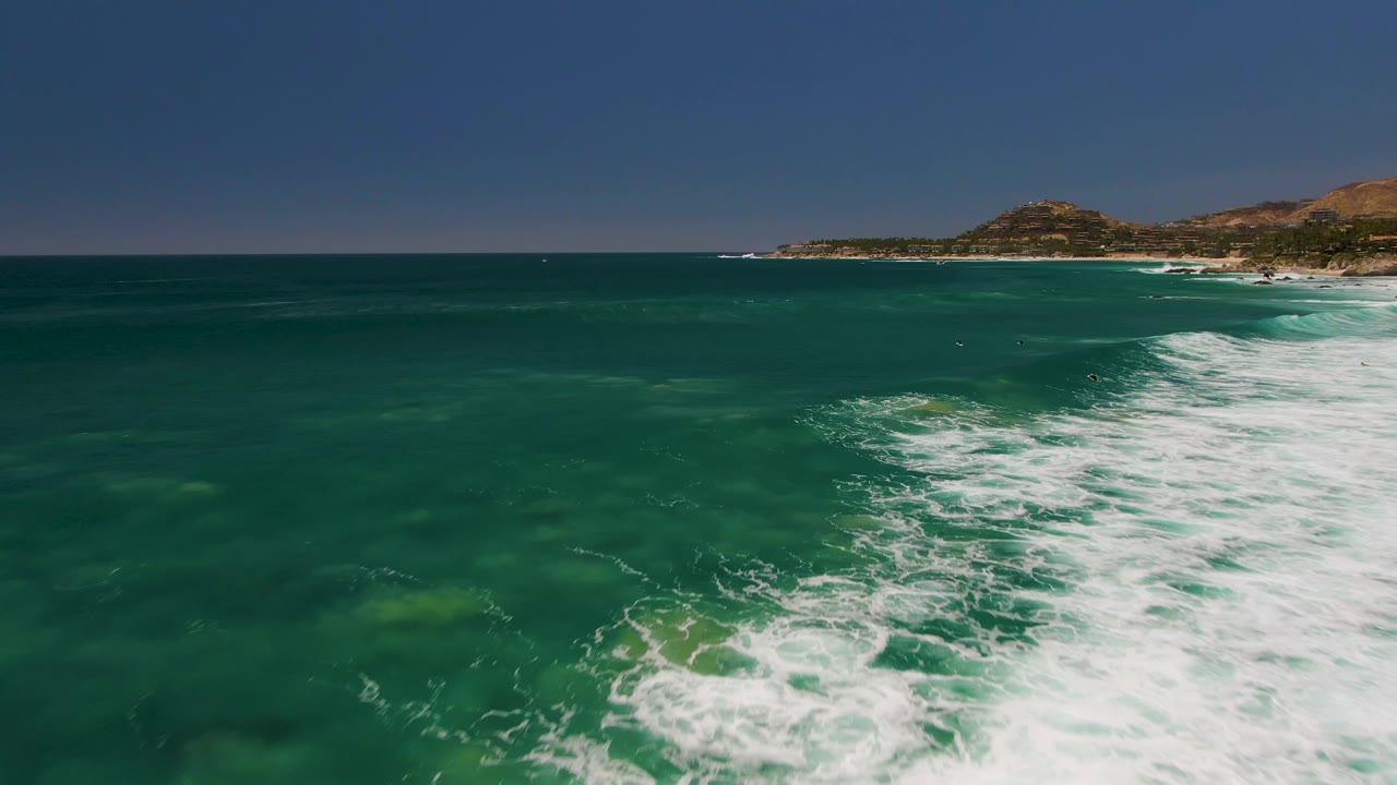 Aerial ocean view at Costa Azul Beach Los Cabos with foamy white water sweeping up the right edge, clearer turquoise water to the left, distant hills, and no visible people.