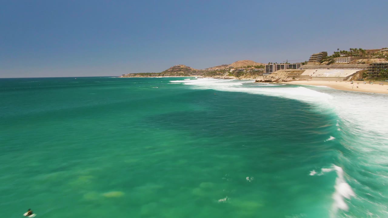 Aerial view of turquoise water and white surf meeting the sandy shore at Costa Azul Beach in Los Cabos, with coastal buildings and hills in the background and no visible people.