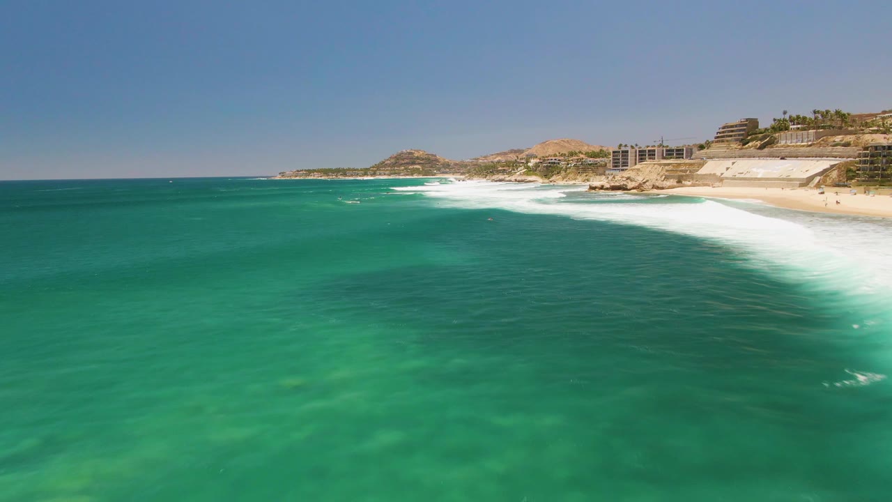 Green-blue ocean water dominates the frame at Costa Azul Beach in Los Cabos while white waves break along the sandy shoreline and low coastal buildings sit beyond the surf.