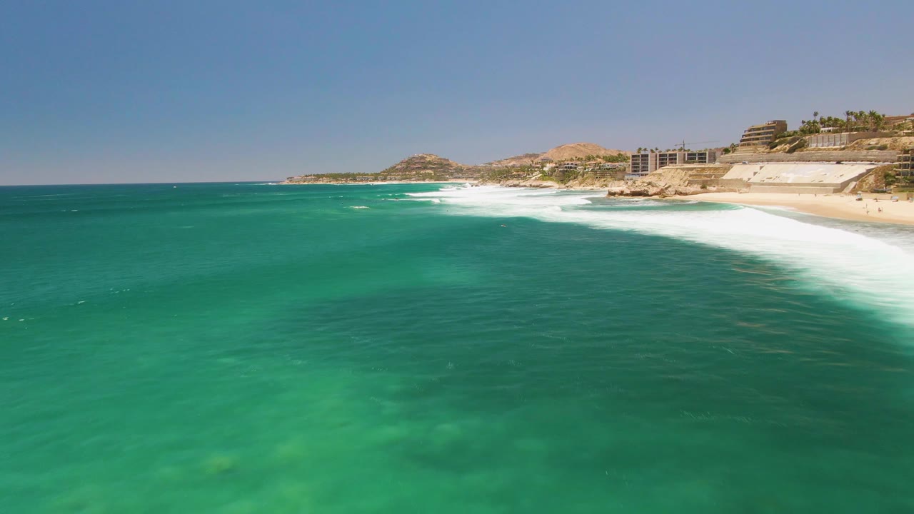 Wide aerial frame over turquoise water at Costa Azul Beach in Los Cabos, with a bright band of white surf, sandy beach, coastal buildings, hills, and no visible people.