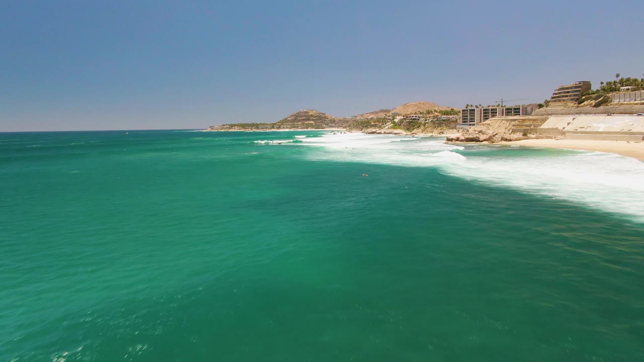 Aerial view at Costa Azul Beach in Los Cabos showing turquoise water in the foreground, white surf breaking near the rocky shoreline, distant beach buildings, low hills, and no visible people.