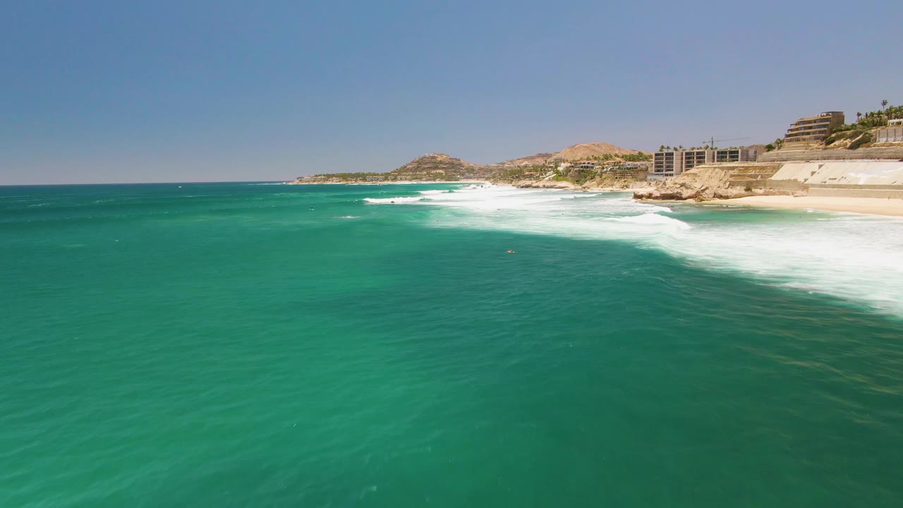 Wide aerial coastal view with turquoise water, breaking waves, beach, rocks, and buildings.