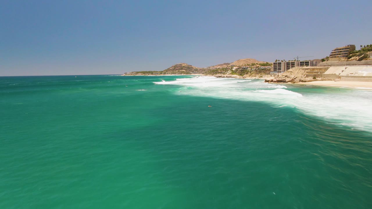 Aerial frame at Costa Azul Beach with a broad patch of white foam near shore, clear turquoise water in the foreground, hillside and buildings in the background, and no visible people.