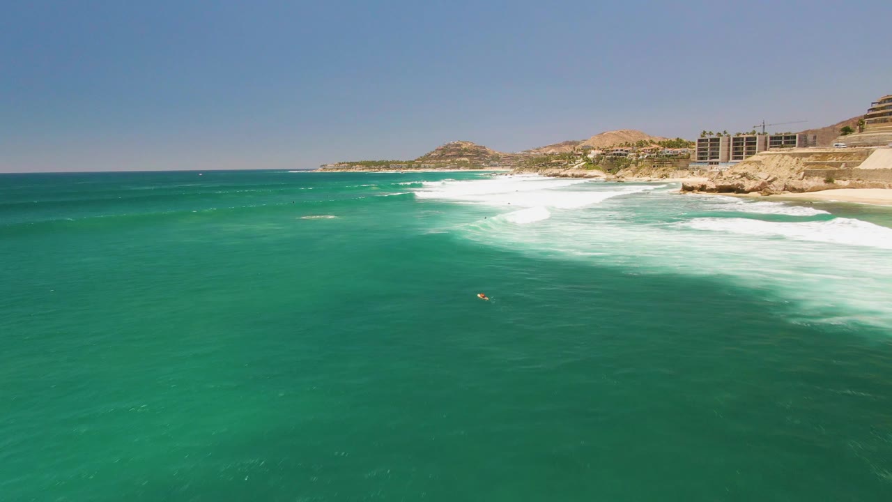 Aerial view over turquoise water at Costa Azul Beach in Los Cabos, with white surf breaking near the rocky shoreline and a tiny swimmer visible offshore.
