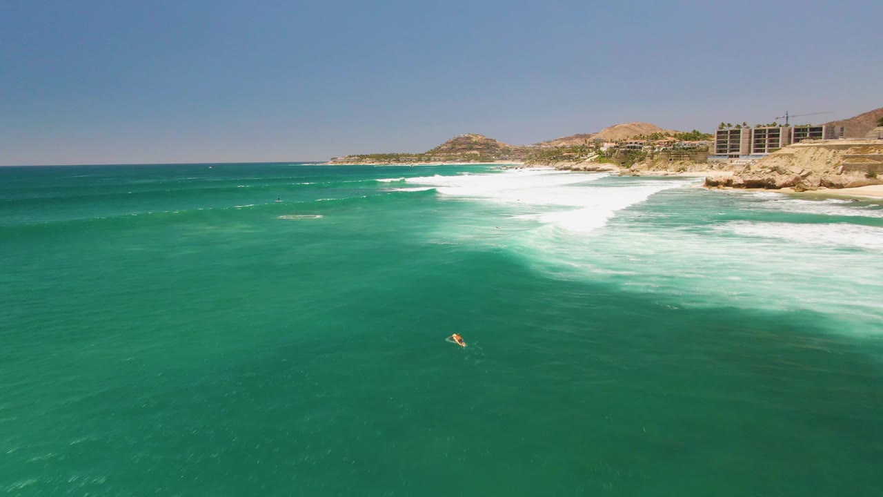 Aerial view of Costa Azul Beach with the swimmer small in the lower right water, white foam spread near shore, and buildings along the rocky coastline.