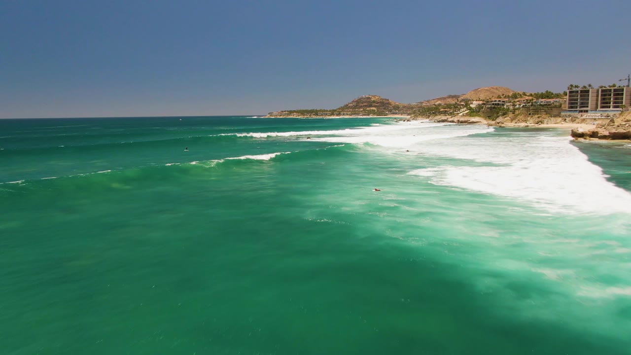 Aerial view of turquoise water and white surf at Costa Azul Beach, Los Cabos, with rocky shoreline, hills, and coastal structures in the distance.