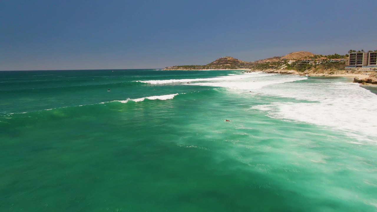 Aerial frame over Costa Azul Beach showing a wide band of white foam crossing turquoise water, with hills and shoreline buildings along the horizon.