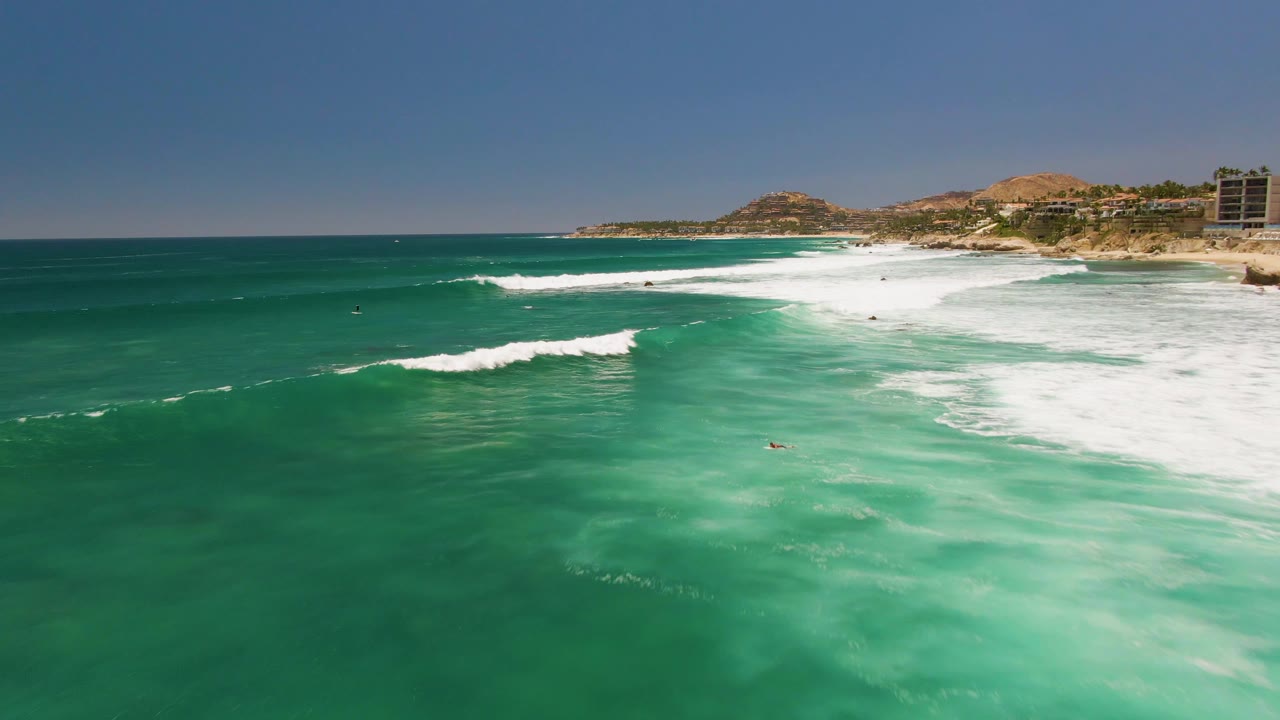 Costa Azul Beach surf seen from above, with layered white wave bands, green-blue water, a rocky point, hills, and structures at the right edge.