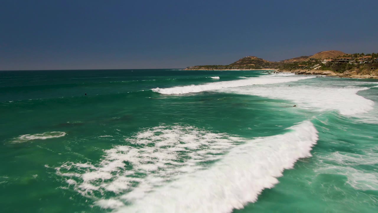 Aerial view at Costa Azul Beach in Los Cabos showing a wide band of white surf breaking across turquoise water, with tiny people visible offshore and hills behind the shoreline.