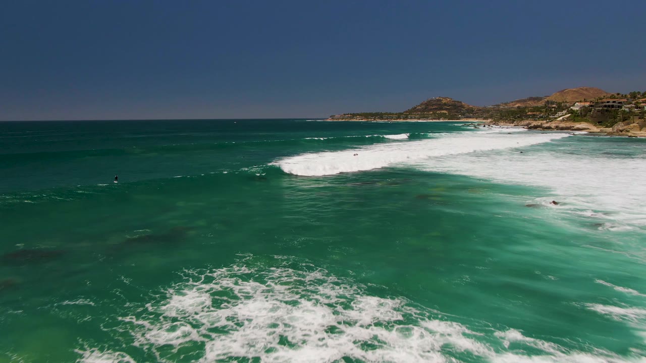 Aerial view over Costa Azul Beach with a broad foamy wave breaking from right to center, scattered people in the water, and the coastline and low hills along the upper frame.