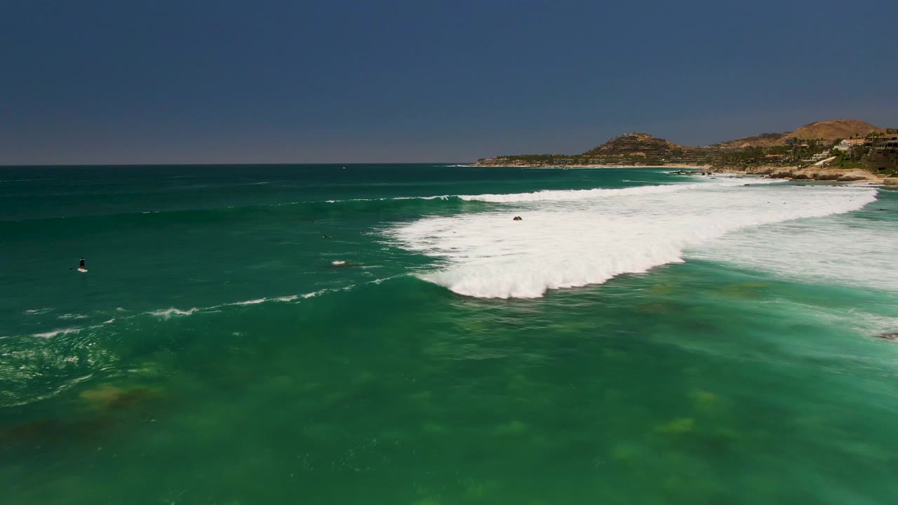 Aerial frame from Costa Azul Beach Los Cabos showing turquoise water, broad white surf, low hills on the coast, and small distant swimmers or surfers in the water.
