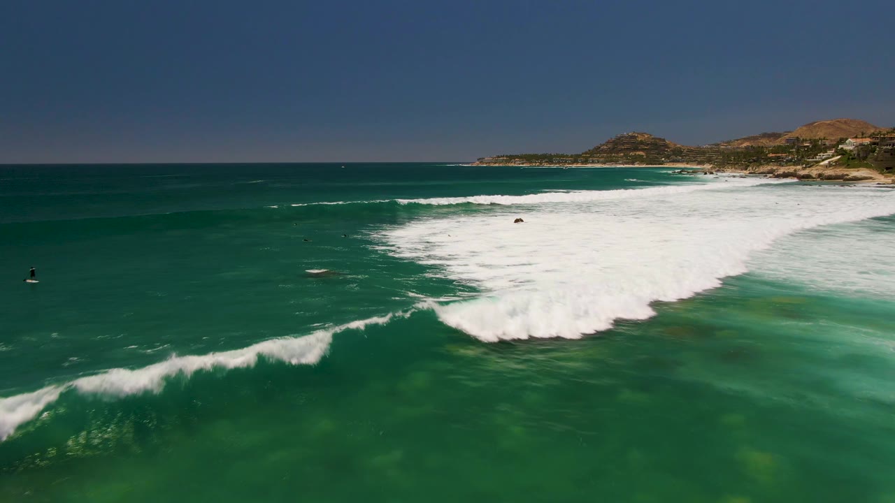 At 0:01, Costa Azul Beach surf pushes farther across the turquoise shallows, with dark distant water figures and a low coastal hillside beyond the wave break.