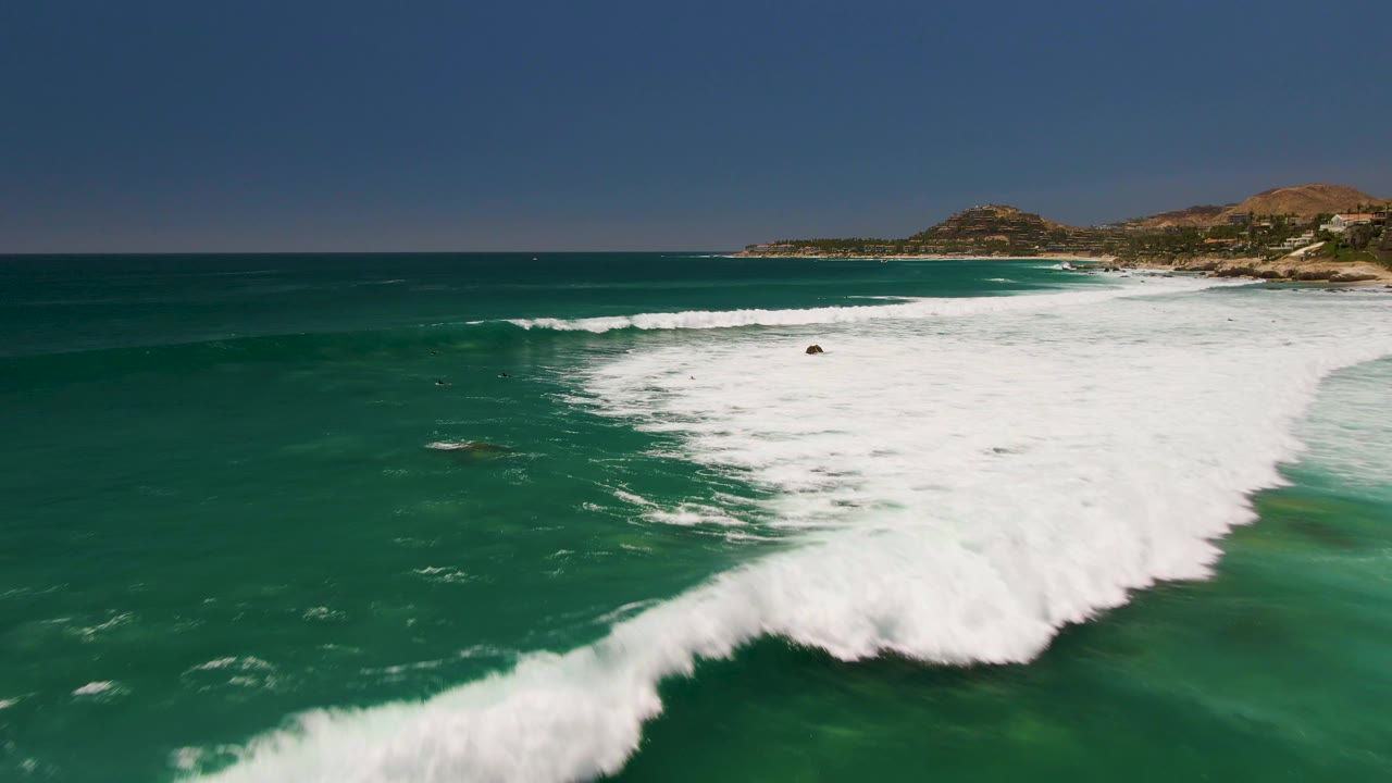 At 0:02, a larger whitewater section fills the right side of the Costa Azul Beach frame while darker open water and small distant swimmers or surfers remain to the left.