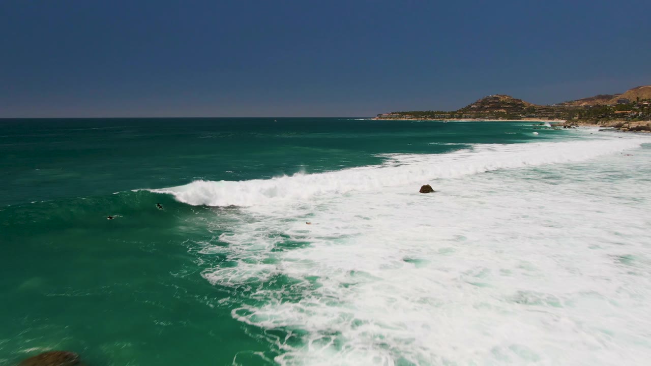 Aerial frame over Costa Azul Beach in Los Cabos showing turquoise water, white surf breaking diagonally, a small offshore rock, distant hills, and tiny people or surfers in the water.