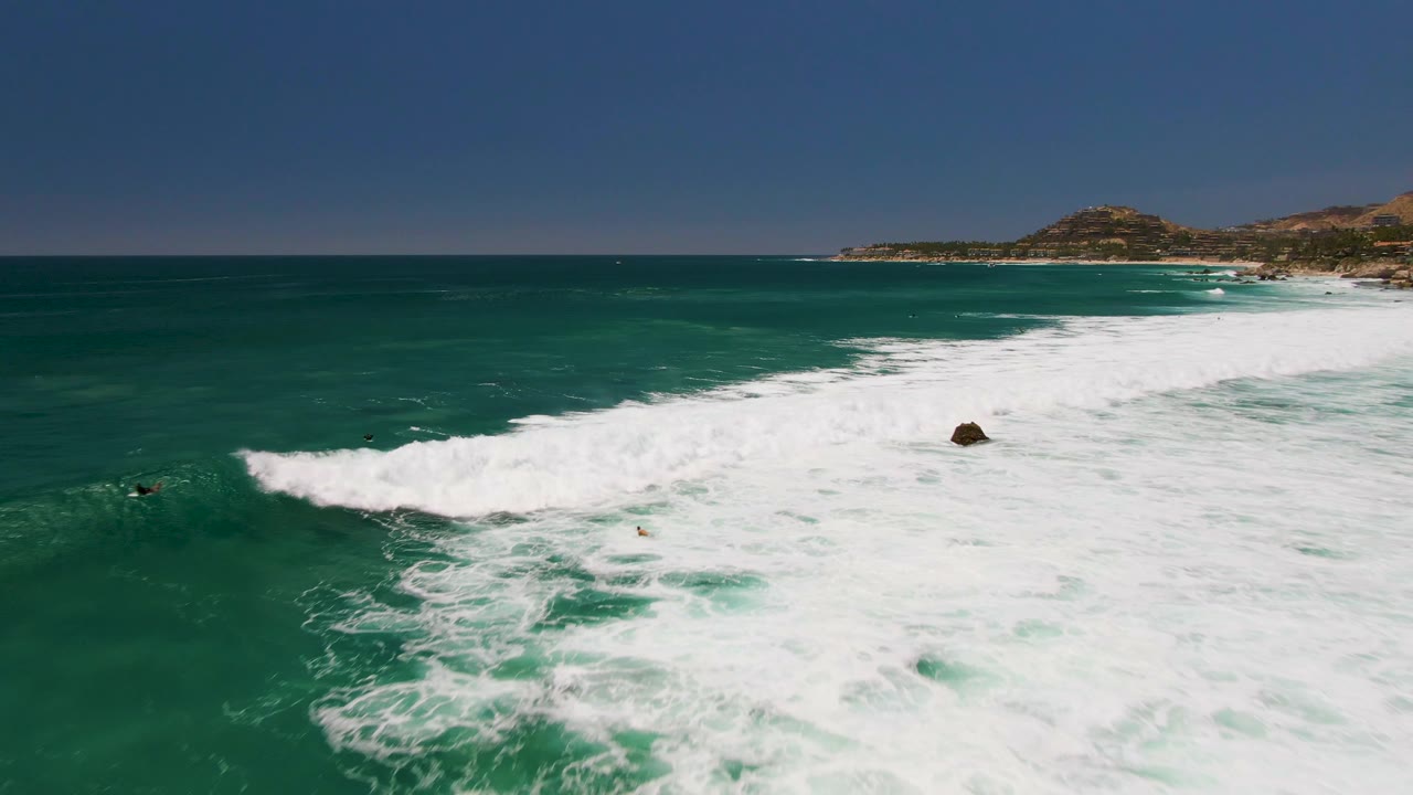 Aerial view at Costa Azul Beach with a wide white breaker spreading across the middle of the frame, dark green water in front, coastline and hills beyond, and tiny distant people or surfers visible.