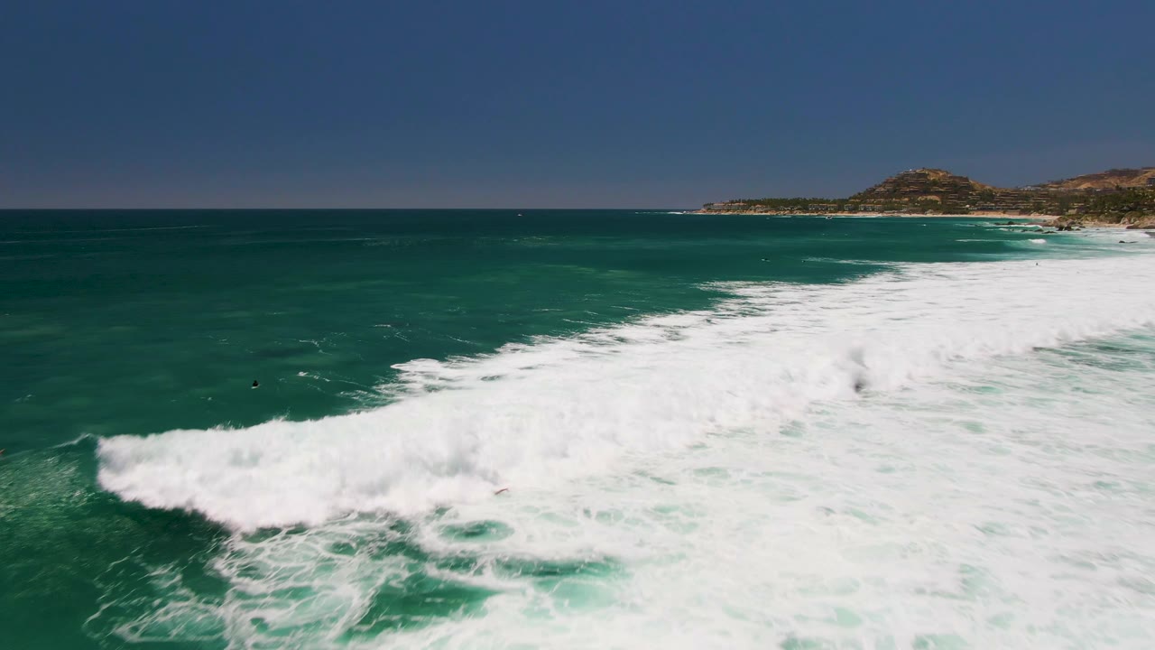 Aerial frame over Costa Azul Beach showing a thicker wall of white surf rolling through green-blue water, with the coastline and hills higher in the background and tiny distant people or surfers offshore.
