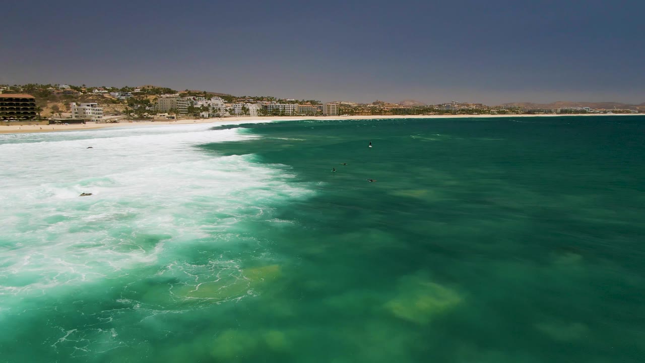 Aerial view at Costa Azul Beach in Los Cabos with white surf filling the left side, green ocean water to the right, small people offshore, and coastal buildings along the sand.