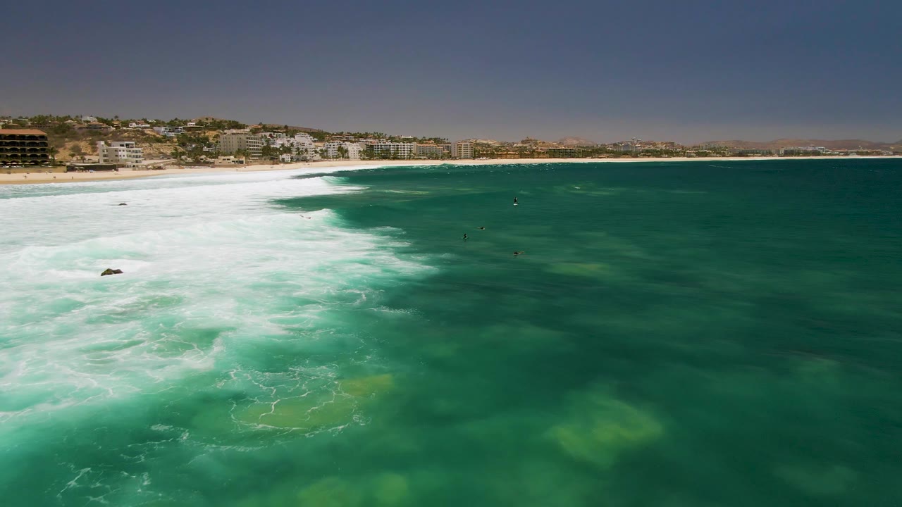 Foamy waves spread across turquoise-green water near a beach backed by buildings.