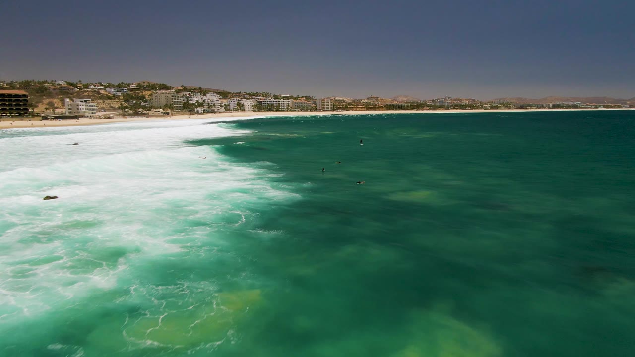At 0:02, the aerial view over Costa Azul Beach shows whitewater pushing diagonally into green sea, scattered small people in the water, and low buildings along the sandy shore.