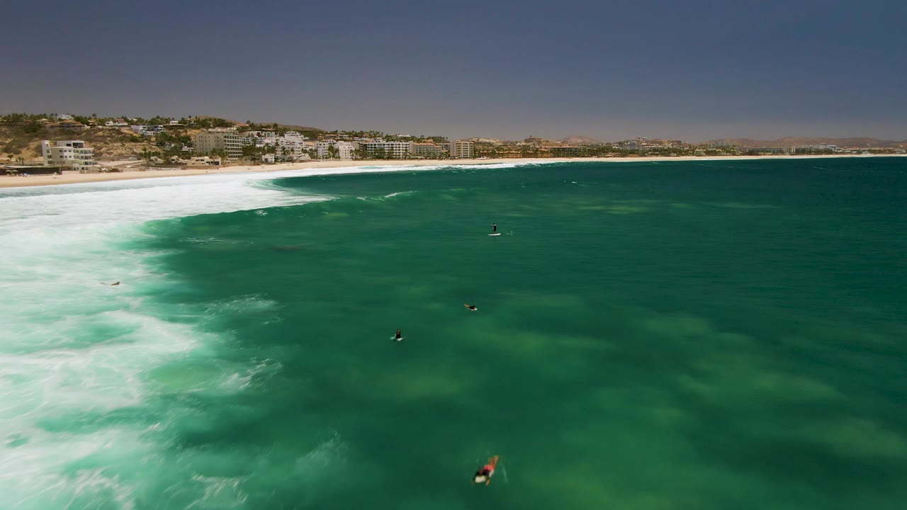 Aerial view of Costa Azul Beach in Los Cabos, with white surf spreading across green water and several distant surfers visible offshore.