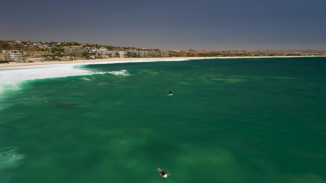 Aerial frame over Costa Azul Beach shows a broad field of green ocean, a white band of surf near shore, and small surfers positioned across the water.