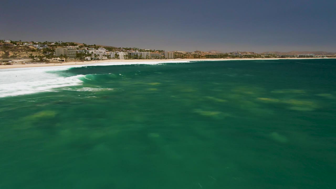 Green water fills the foreground at Costa Azul Beach as white surf reaches the sandy shore and hillside buildings line the background, with no visible people.