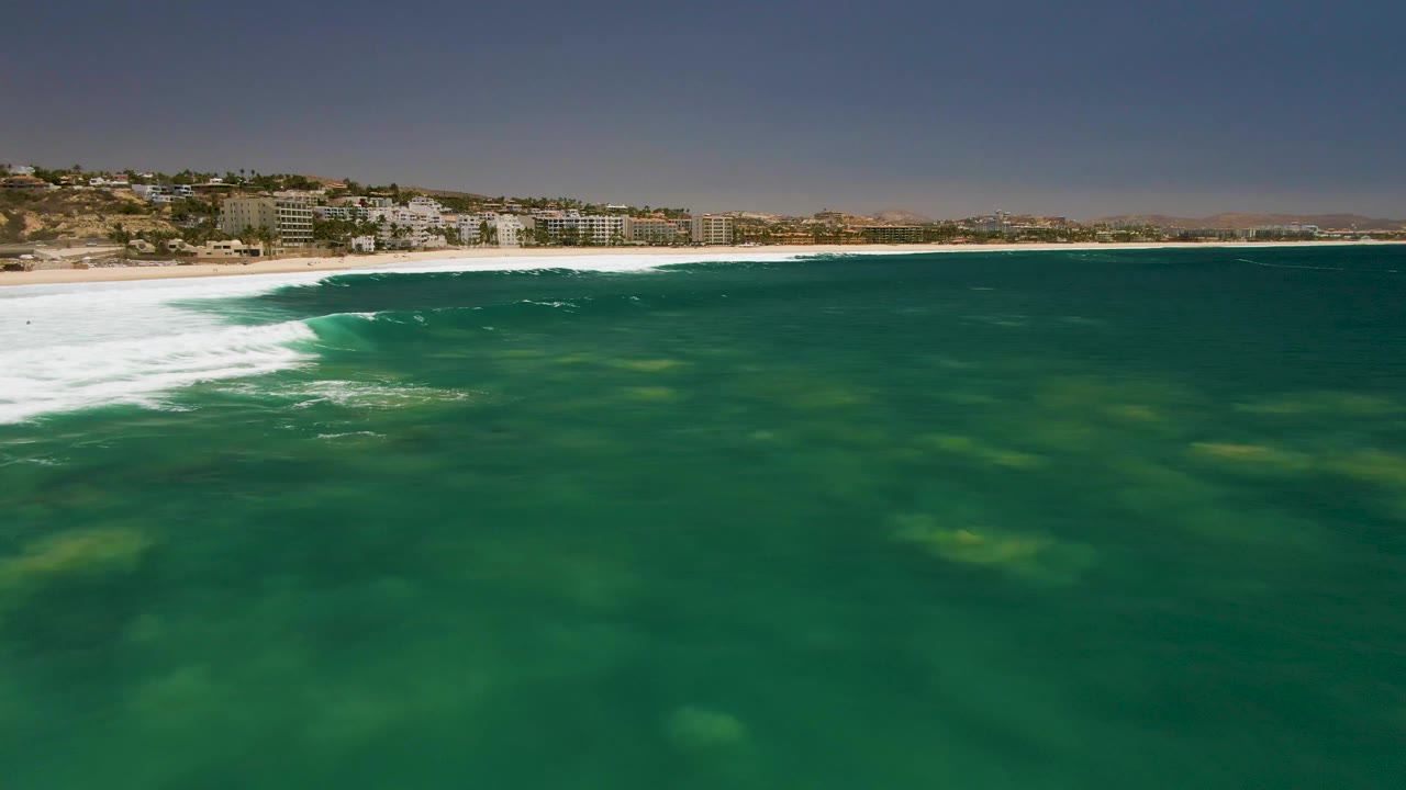At Costa Azul Beach, a band of white foam pushes along the left shoreline while green ocean water covers the foreground and coastal buildings remain in the distance, with no visible people.